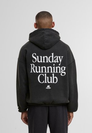 Mann trägt einen schwarzen Kapuzenpullover mit dem Text "Sunday Running Club" in Weiß und steht, mit dem Rücken zur Kamera, vor einem schlichten grauen Hintergrund.