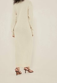 Vestito lungo in maglia color crema con maniche e orlo a coste, caratterizzato da una silhouette ampia e da un tessuto testurizzato. Abbinato a sandali con tacco marroni.