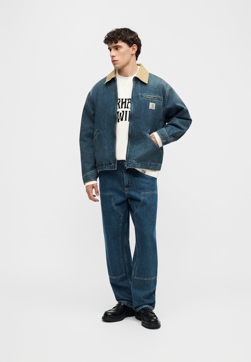 Giovane uomo che indossa una giacca di jeans con colletto marrone chiaro, felpa bianca con motivo grafico, jeans coordinati e scarpe nere, in piedi con una mano in tasca.