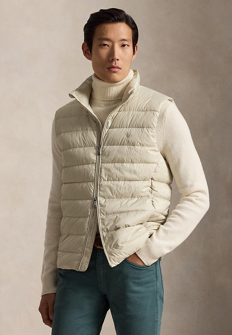 Homme portant une veste matelassée beige sur un pull à col roulé crème avec un pantalon bleu sarcelle, debout avec une main dans la poche devant un fond uni.