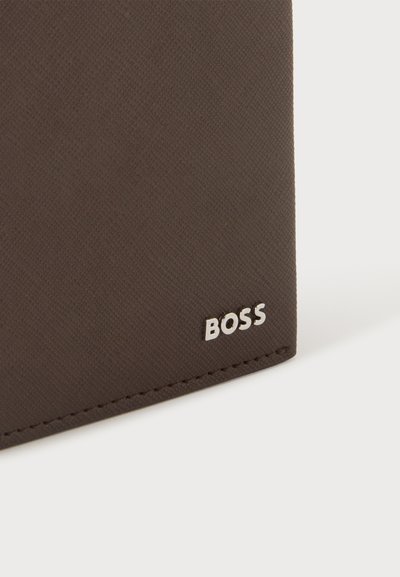 Gros plan sur le coin d'un portefeuille texturé marron foncé avec un petit logo métallique argenté "BOSS" et des coutures visibles sur fond clair.