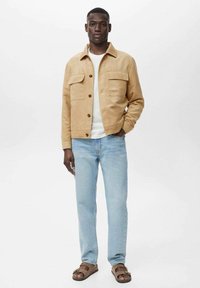 Tan suede jacket with two chest pockets, worn over a white shirt, paired with light blue jeans and brown sandals. Plain white background.