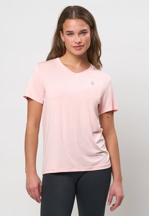 Jauna sieviete ar gariem brūniem matiem, tērpusies gaiši rozā V veida kakla īsām piedurknēm kreklā un melnās legingās, stāv pret vienkāršu fonu.