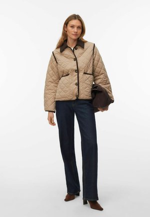 Femme portant une veste matelassée beige avec des bordures foncées, un jean bleu foncé et des bottes à talons brunes, tenant un manteau foncé plié dans sa main gauche.
