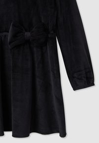 Abito in corduroy nero con maniche lunghe, caratterizzato da un grande fiocco in vita e texture a righe verticali. Dettaglio di polsini rifiniti.