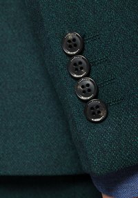 Ciemnozielony materiał marynarki z teksturowanym wzorem. Cztery czarne guziki z czterema otworami, ułożone pionowo na mankiecie rękawa.