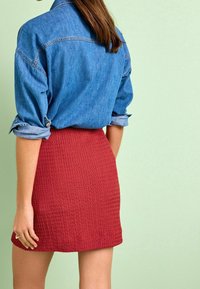 Camicia di denim con maniche arrotolate, abbinata a una gonna mini rossa e strutturata. Stile semplice e casual con una finitura liscia e colori a contrasto.