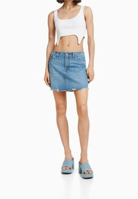 Donna che indossa un top bianco corto, una mini gonna di jeans blu scucita e sandali open-toe con piattaforma azzurri, in piedi contro uno sfondo bianco.