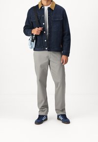 Tumma farkkutakki, jossa on vaaleanruskea corduroy-kaulus, harmaat housut ja siniset tennarit kolmella raidalla. Pitää kädessään vaaleansinistä olkalaukku.