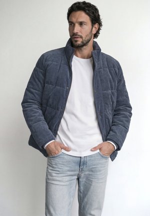 Homme portant une veste matelassée bleue sur une chemise blanche et un jean bleu clair, debout les mains dans les poches, regardant sur le côté.