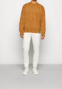 Ruskeakuvioinen collegepaita pitkällä hihalla ja pyöreällä kaula-aukollla, yhdistettynä valkoisiin housuihin ja valkoisiin tennareihin harmaalla taustalla.