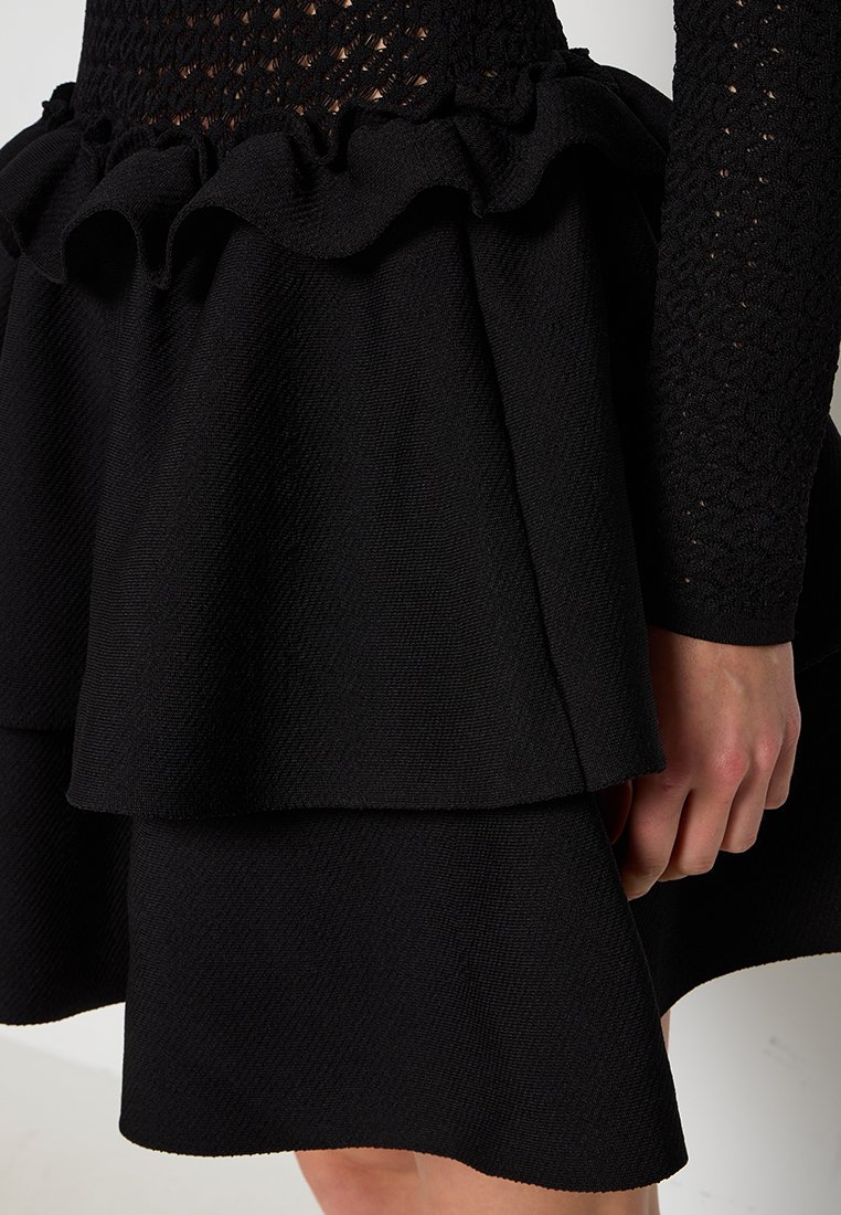 Schwarzes Kleid mit einem strukturierten, taillierten Oberteil, langärmeligem Strickdesign und einem gestuften, rüschigen Rock mit glatter Oberfläche.