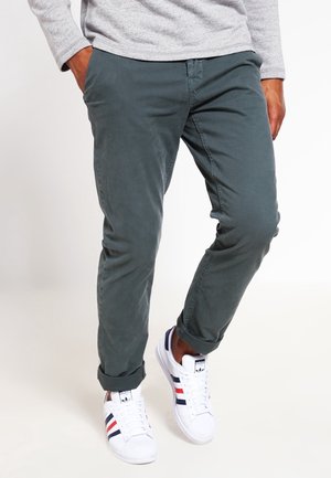 Man draagt opgerolde donkergroene broek, een lichtgrijs shirt met lange mouwen en witte Adidas sneakers met rode en marineblauwe strepen.