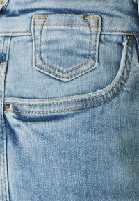 Světle modrá džínovina s malou zadní kapsou s špičatým klopou, zvýrazněná viditelným šitím a mírně třepícím se okrajem.