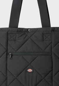 Svart quiltet totebag med lomme foran med glidelås, trykknaplukking og brede stoffhåndtak; liten rektangulær logopatch plassert sentrert under glidelåsen.