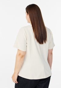 Beżowa bluzka z krótkim rękawem o gładkiej fakturze, dopasowana sylwetka i zaokrąglony dół, zestawiona z ciemnymi dżinsami.