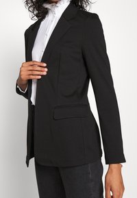 Nainen pukeutuneena mustaan räätälöityyn bleiseriin, jonka alla on valkoinen volangikauluspaita ja tummat farkut. Hän koskettaa toisen käden sormin bleiserin lievettä.