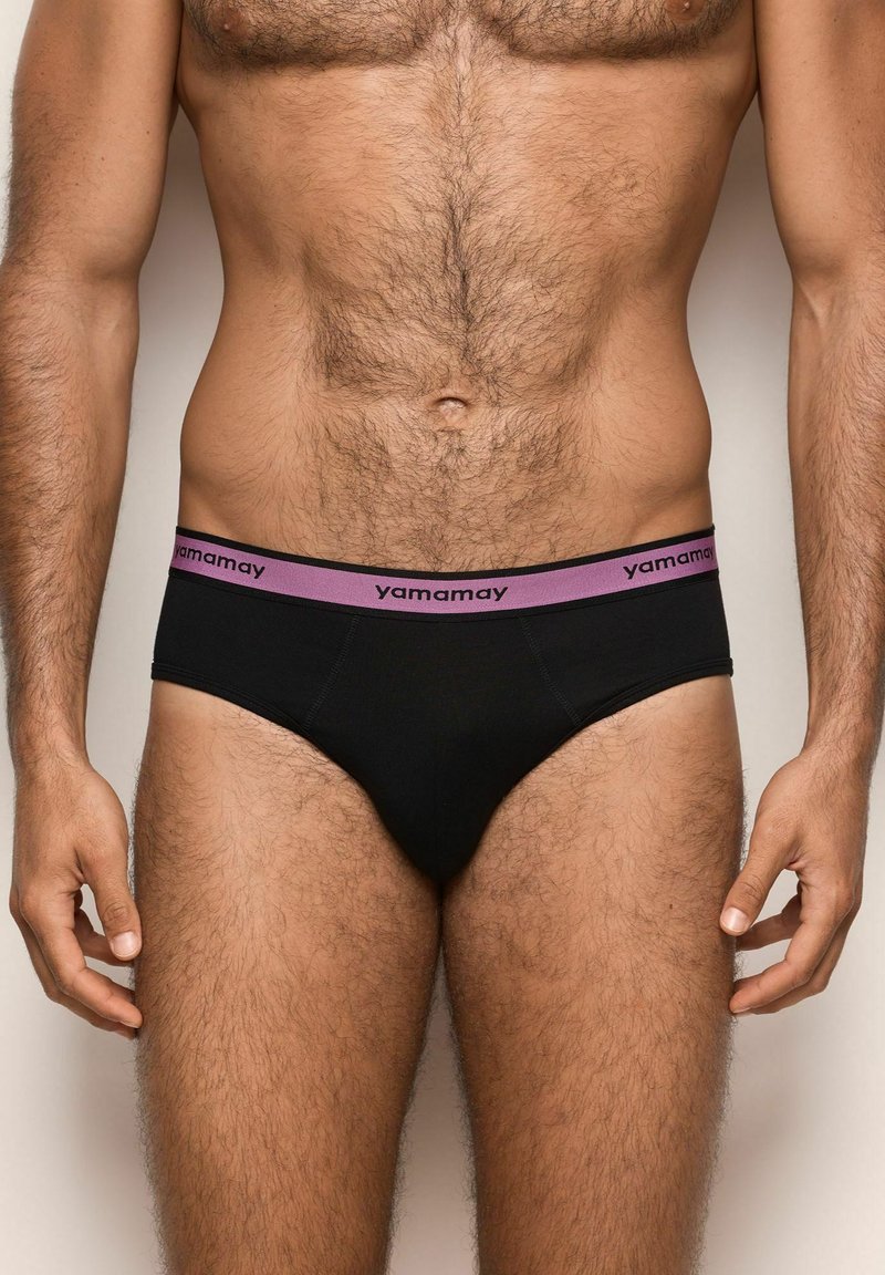 Männlicher Oberkörper, der schwarze Unterhosen mit einem lila Bündchen trägt, auf dem der Markenname "yamamay" auf einem schlichten Hintergrund zu sehen ist.