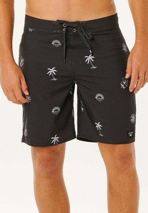 Pantalones cortos de baño negros con patrones blancos de palmeras, sol y flores, usados por una persona de pie mostrada de la cintura a las rodillas.