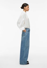 Blouse blanche légère à manches bouffantes associée à un jean bleu en denim taille haute à jambes droites. Des chaussures à talons foncées complètent le look.