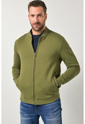 Homme portant une veste zippée vert olive sur un t-shirt foncé, les mains dans les poches, accompagné d'un jean bleu, faisant face à l'avant sur un fond uni.