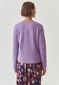 Lila gestrickter Pullover mit weicher Textur, lockerer Passform und Rippdetails am Saum. Getragen über einem bunten gemusterten Rock.