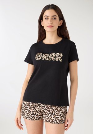 Donna che indossa una maglietta nera con la scritta "GRRR" a lettere leopardate e pantaloncini abbinati con stampa leopardata, in piedi davanti a uno sfondo bianco.