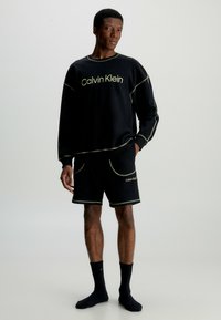 Černá souprava mikiny a šortek s kontrastním švem; obě položky mají zelené logo "Calvin Klein"; obyčejné černé ponožky.