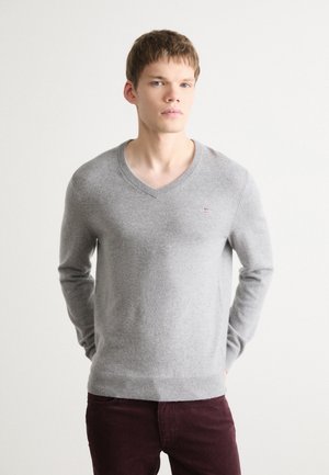 Suéter gris con escote en V, mangas largas y puños acanalados. Textura suave, detalle sutil del logo en el pecho. Combinado con pantalones en un marrón oscuro.
