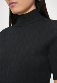 Crni pleteni džemper s visokim rebrastim ovratnikom i teksturiranim uzorkom dijamanta, prikazan izbliza radi naglašavanja detalja tkanine.