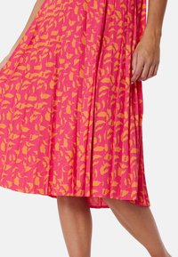 Roze geplooide jurk met een oranje bladerenpatroon, gemaakt van een lichte stof met een zachte textuur. De zoom valt boven de knie.