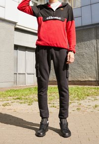 Sudadera con capucha de forro polar en rojo y negro con cremallera de medio cierre y logo, combinada con pantalones cargo negros con bolsillos laterales y zapatillas negras.