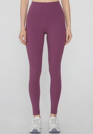 Lila hochtaillierte Leggings mit glatter Textur. Sie haben ein körperbetontes Design, das bis zu den Knöcheln reicht. Kombiniert mit weißen Sportschuhen.