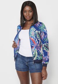 Giacca bomber con uno sfondo blu e motivi di foglie tropicali in rosa e verde, con polsini a coste e zip frontale.