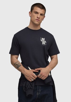 Joven con el pelo corto que lleva una camiseta gráfica negra, sosteniendo una chaqueta negra alrededor de la cintura, con un tatuaje y anillos visibles.