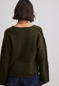 Vrouw met kort bruin haar, gekleed in een losse donkergroene gebreide trui en een spijkerbroek, gezien vanachter tegen een effen lichtachtergrond.