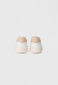 Zapatilla blanca y beige con parte superior texturizada, talón acolchado y gruesa suela blanca. Acabado suave con sutiles acentos y forma redondeada.