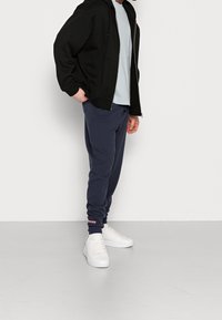 Černá mikina na zip, světle modré tričko, tmavě modré tepláky a bílé tenisky. Hladká látka s elastickými manžetami a logo nášivkou na kalhotách.