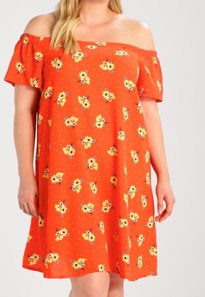 Žena v krátkých, krátkých rukávech šatech s odhalenými rameny v oranžové barvě s žlutým květinovým vzorem, stojící proti jednobarevnému pozadí.