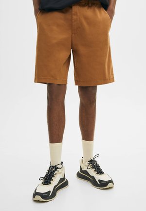 Braune Baumwollshorts mit elastischem Bund, Seiten Taschen, einem schlichten Design, kombiniert mit schwarz-weißen Sneakern und hellen gerippten Socken.