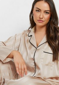 Satinpyjamaset i ljusbeige med subtila ränder, svarta detaljer vid kragen och fickan, samt löst sittande långa ärmar.