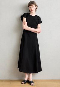 Crna haljina kratkih rukava s okruglim izrezom, lepršavim rubom i suptilnom teksturom. Uparena s ravnim crnim sandalama. Jednostavan, elegantan dizajn.