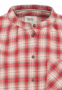 Chemise à carreaux rouges et blancs avec un col officier, deux boutons marron, et une poche poitrine gauche arborant un motif à carreaux assorti.