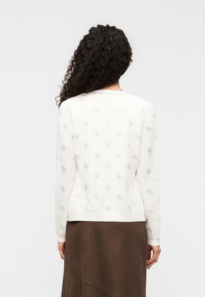 Femme aux cheveux bouclés portant un pull blanc à manches longues avec de petits motifs répétés et une jupe marron, de dos sur un fond uni.