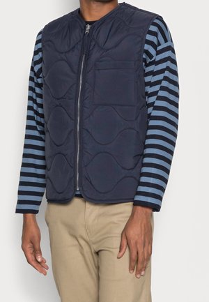 Homme portant un gilet matelassé bleu marine avec fermeture éclair devant, sur une chemise à manches longues rayée bleue et noire, et un pantalon beige, debout sur fond blanc.