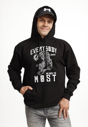 Uśmiechnięty mężczyzna w czarnej bluzie z kapturem z grafiką tygrysa i napisem „WSZYSCY WIEDZĄ, ŻE JESTEŚMY NAJLEPSI”, czarna czapka i szare dżinsy, pozujący nonszalancko.