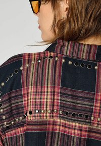 Chemise à carreaux présentant des couleurs rouge, jaune et marine, ornée de clous métalliques et d'accents circulaires le long des coutures des épaules.