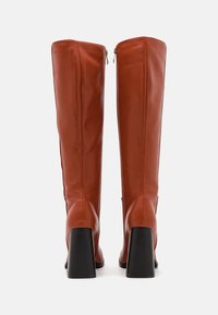 Bottes hautes en cuir marron, droites, à texture lisse, fermeture éclair, talons noirs épais, affichant un design élégant et moderne.