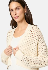 Brede gebreide cardigan in crème met een losse, open structuur, voorzien van geribbelde manchetten en zoom. Model draagt een witte top eronder.