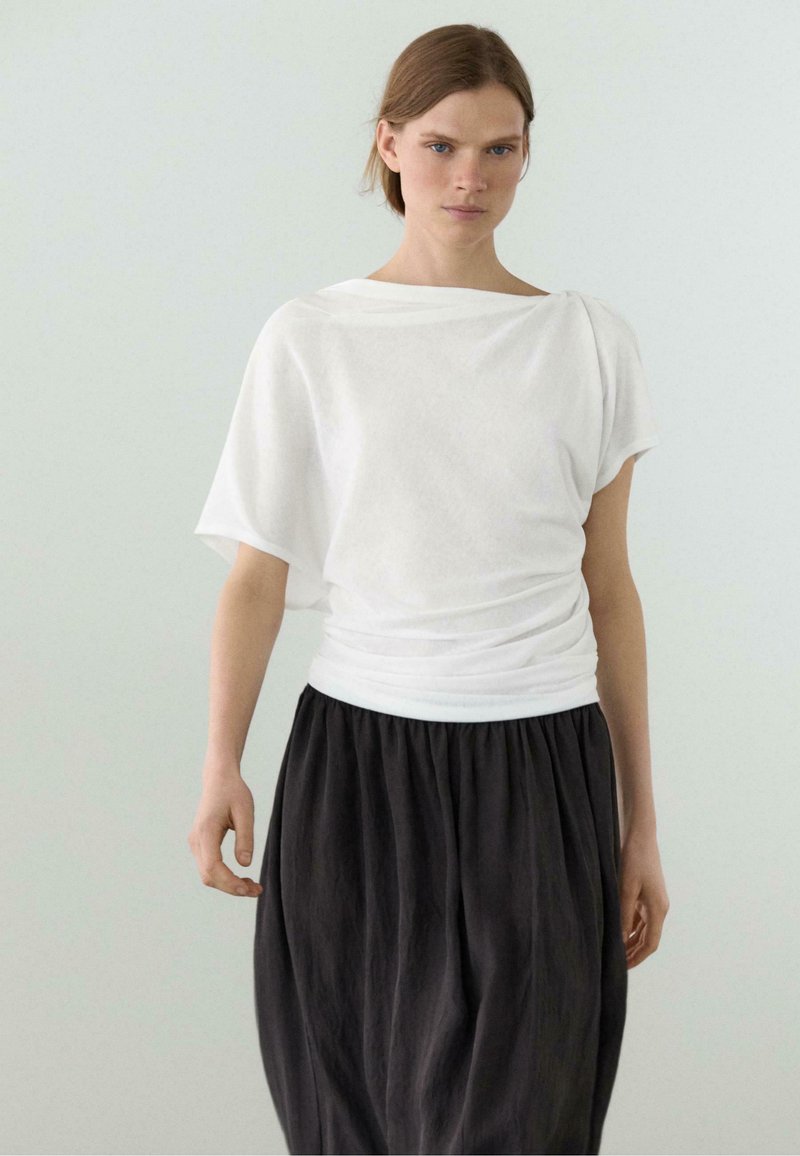 Vrouw met licht haar die een losse witte top en een hoge taille, geplooide zwarte rok draagt, staand tegen een eenvoudige lichte achtergrond.